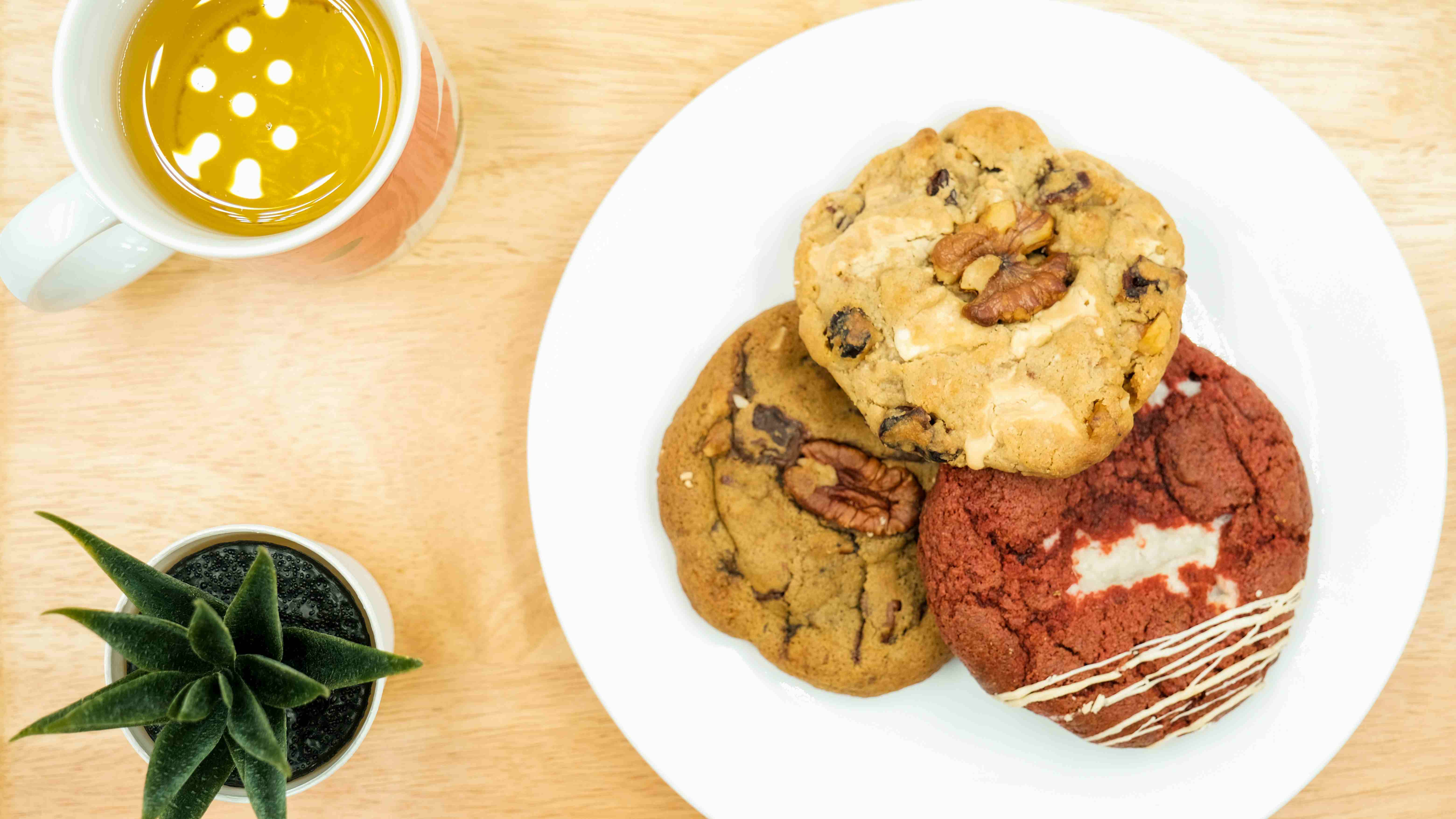 Tre biscotti artigianali su un piatto bianco: uno con noci pecan e gocce di cioccolato, uno con cioccolato bianco e frutta secca, e uno rosso tipo red velvet con decorazioni di glassa. Accanto, una tazza di tè e una piantina su tavolo in legno chiaro.